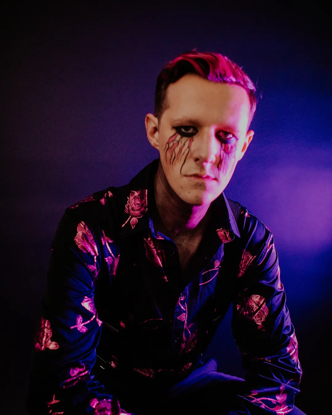 Kyle Kupczyk seated in a studio portrait, lit in pink and blue, wearing a black rose-print shirt