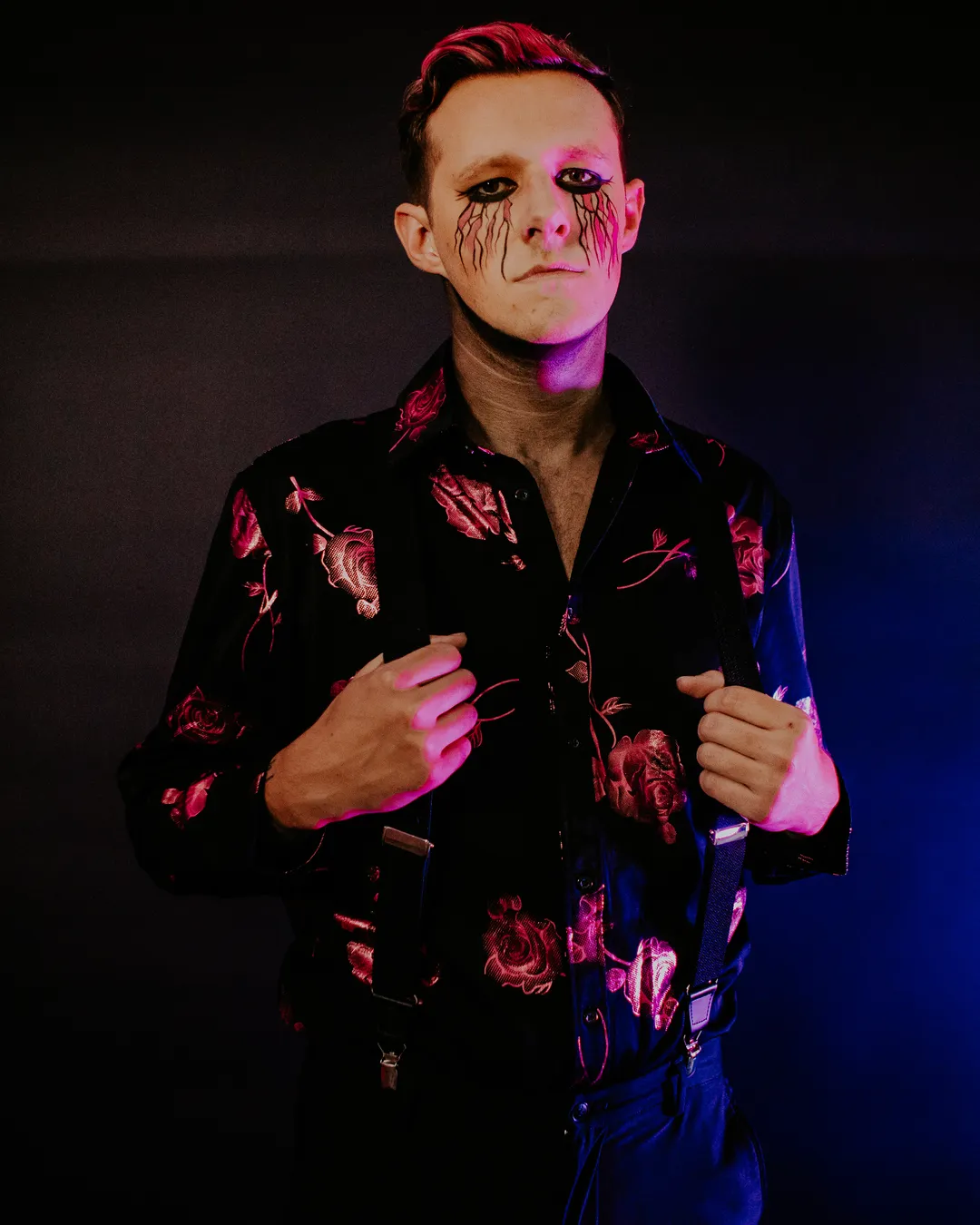 Kyle Kupczyk standing in a rose-print shirt and suspenders against a dark backdrop
